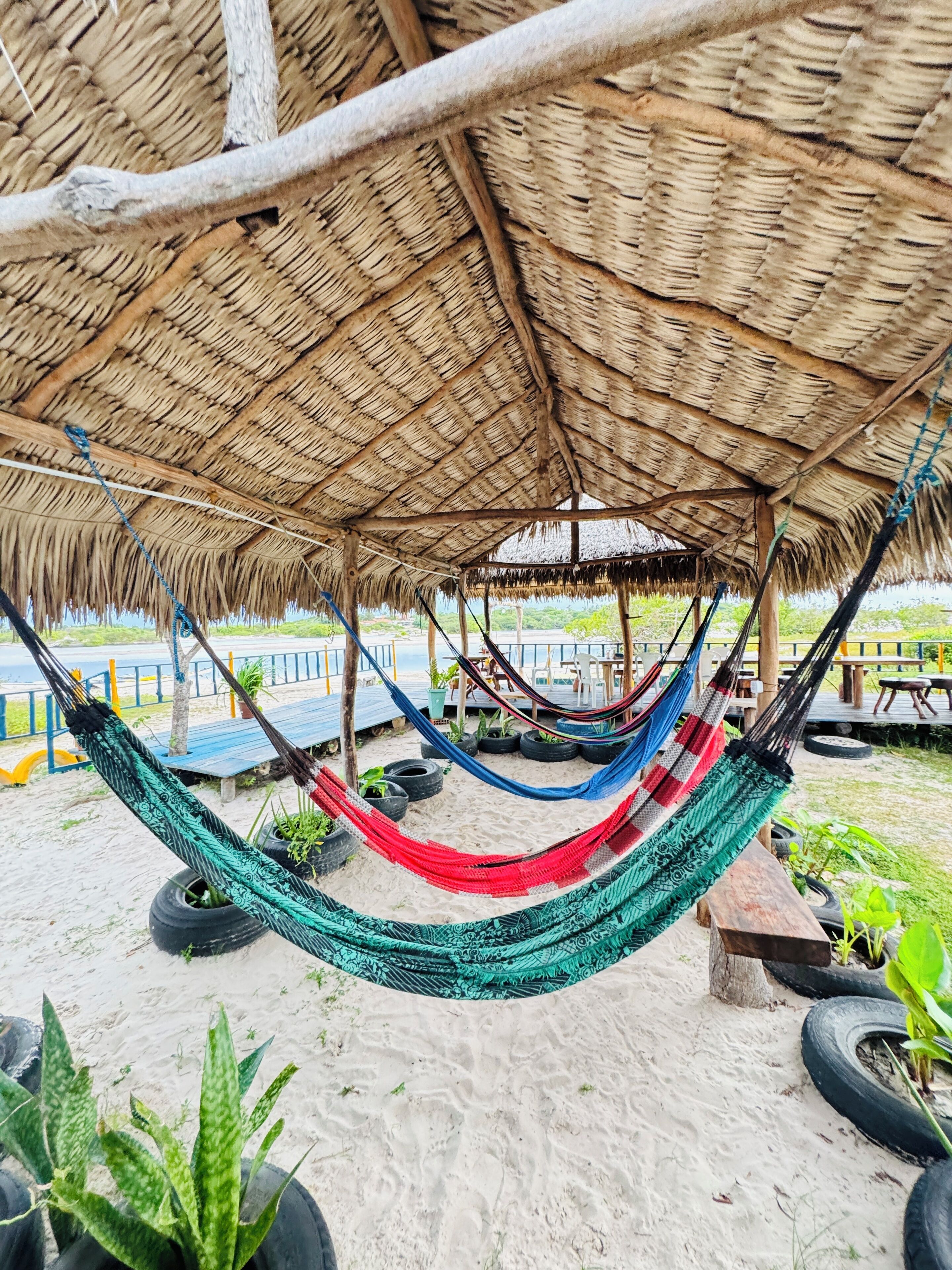 A hammock under a thatched roof structure, with a sandy ground and tire planters.