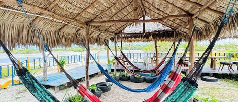 A hammock under a thatched roof structure, with a sandy ground and tire planters.
