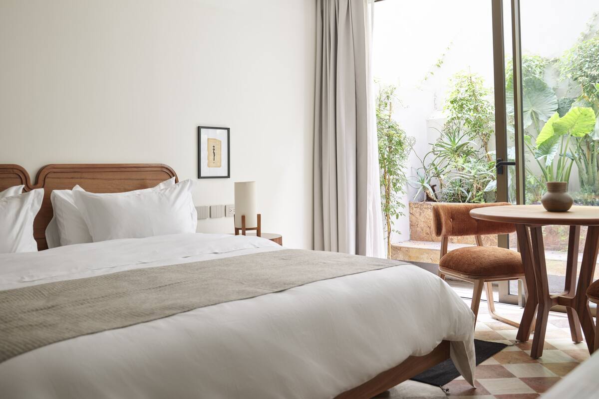 A hotel room with a large bed, a small table with a vase, and a view of greenery outside.