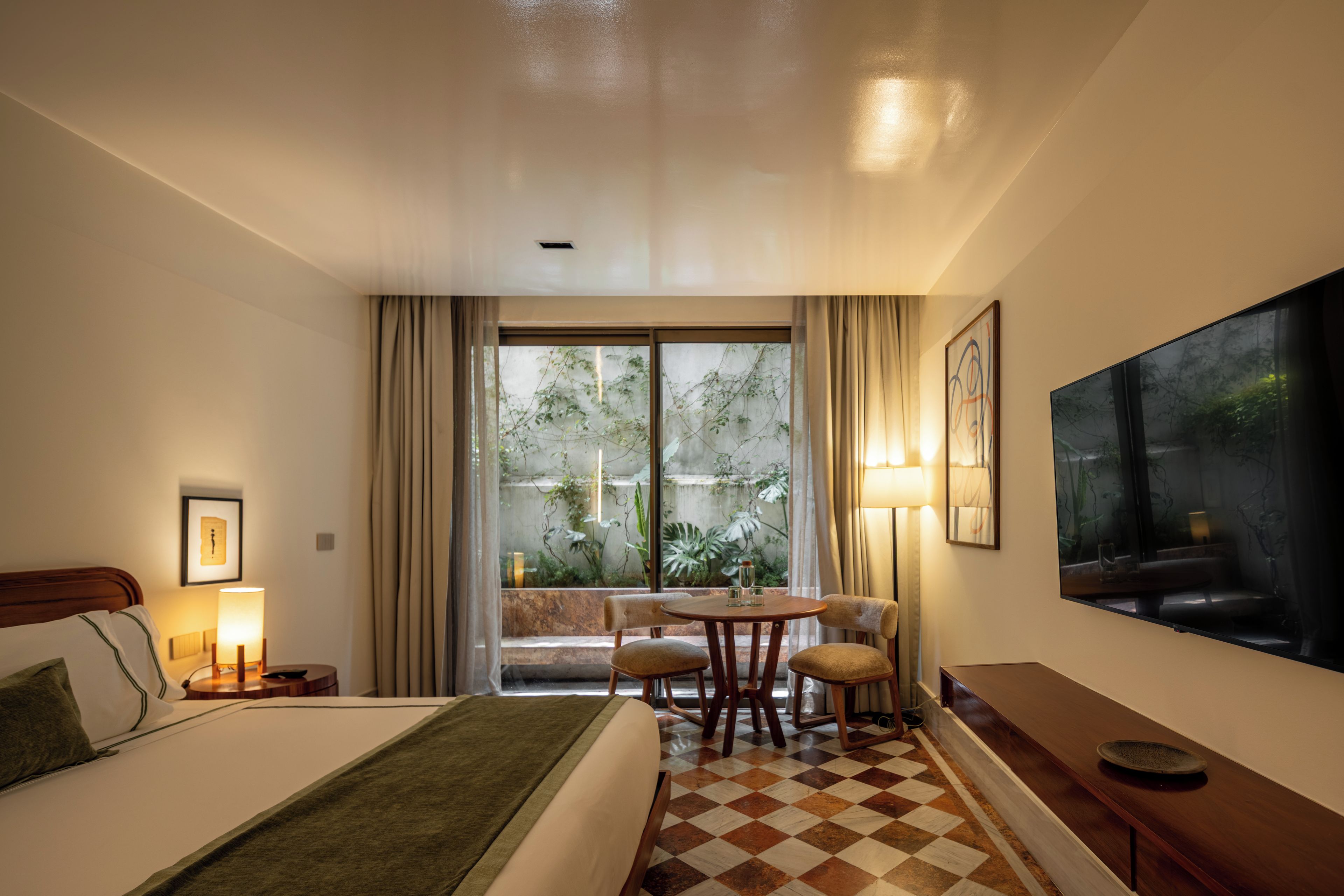 Habitación de hotel con cama, una mesa de comedor con sillas, un ventanal con cortinas y un cuadro en la pared.