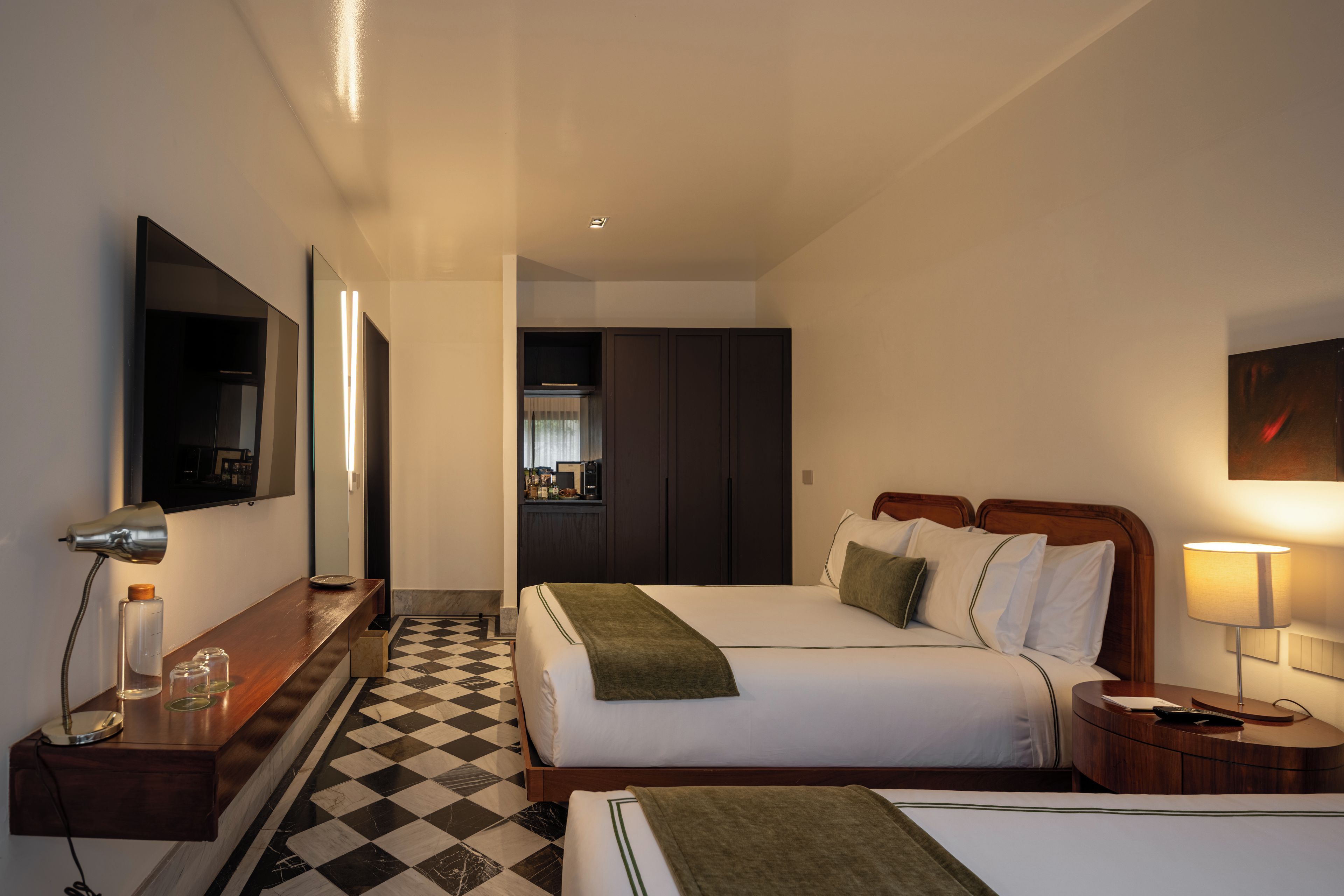 A hotel room with a checkered floor, a bed with pillows, a bedside table with a lamp, a mirror, and a television.