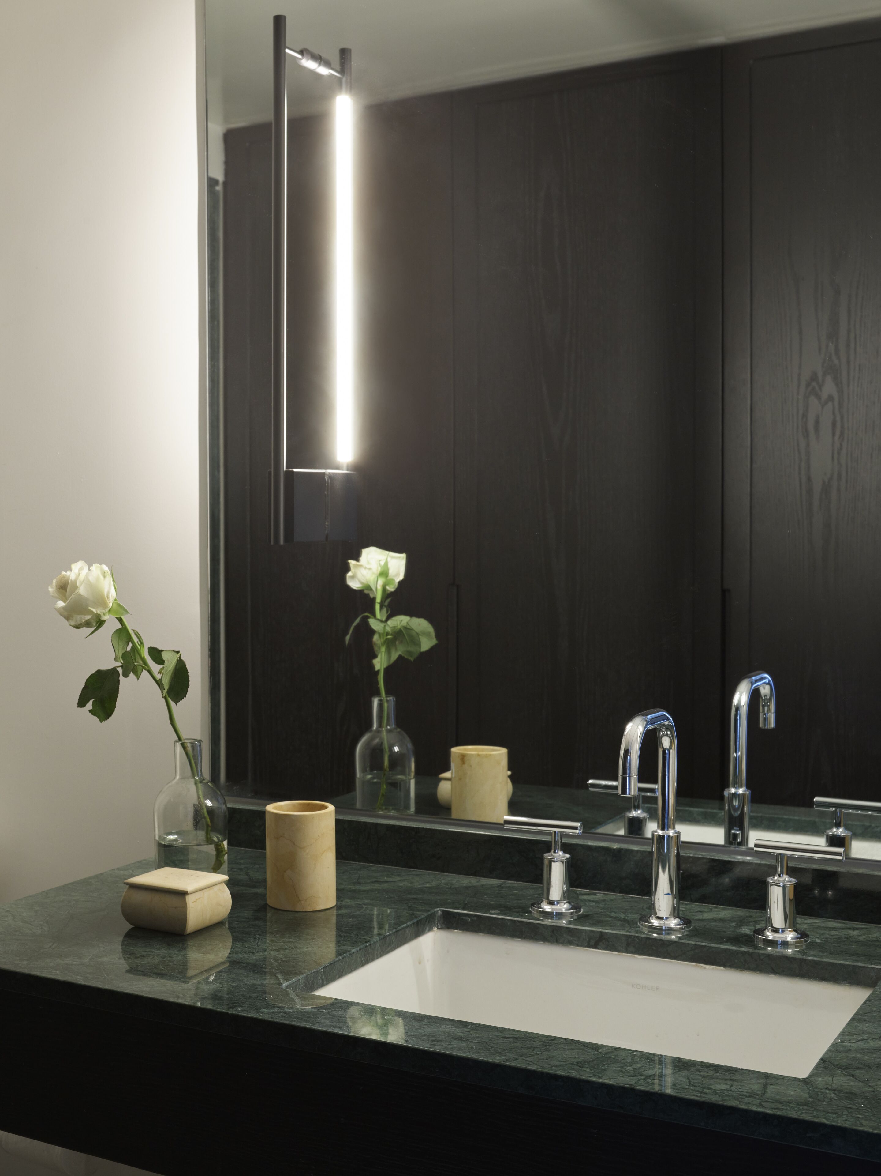 A bathroom with a dark wood vanity, a white sink, two faucets, and a mirror reflecting the room. There are two vases with flowers on the vanity.