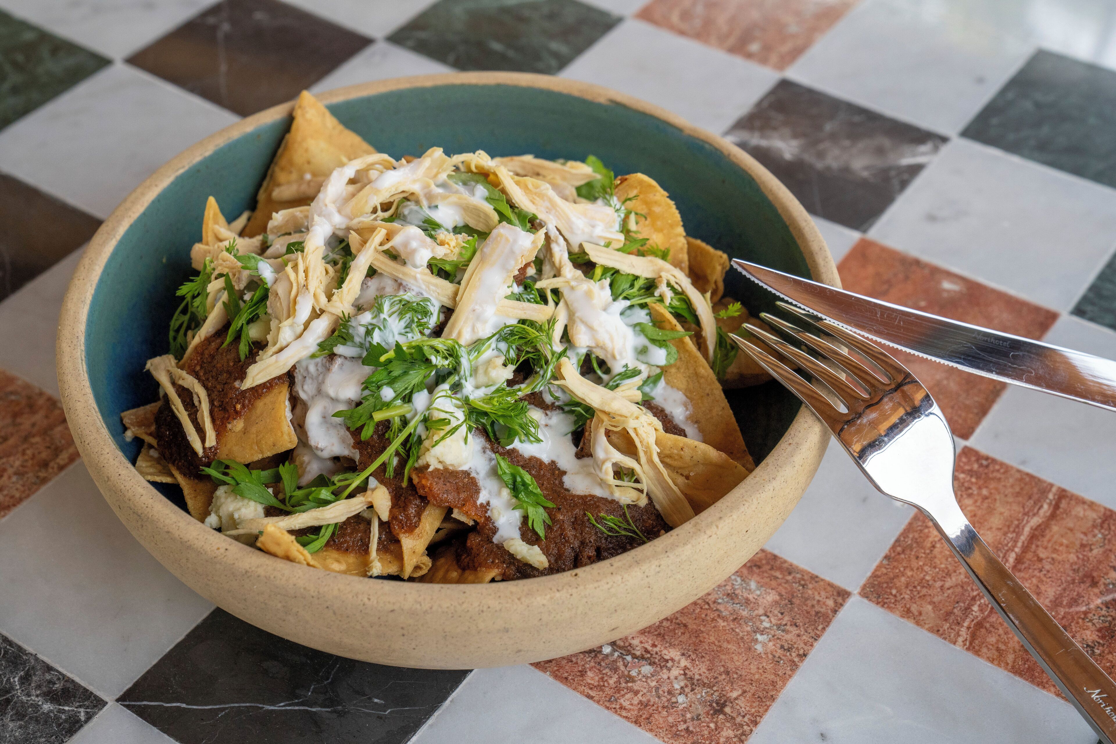 A bowl of nachos topped with shredded chicken, cheese, and a creamy sauce, served on a checkered tile floor.