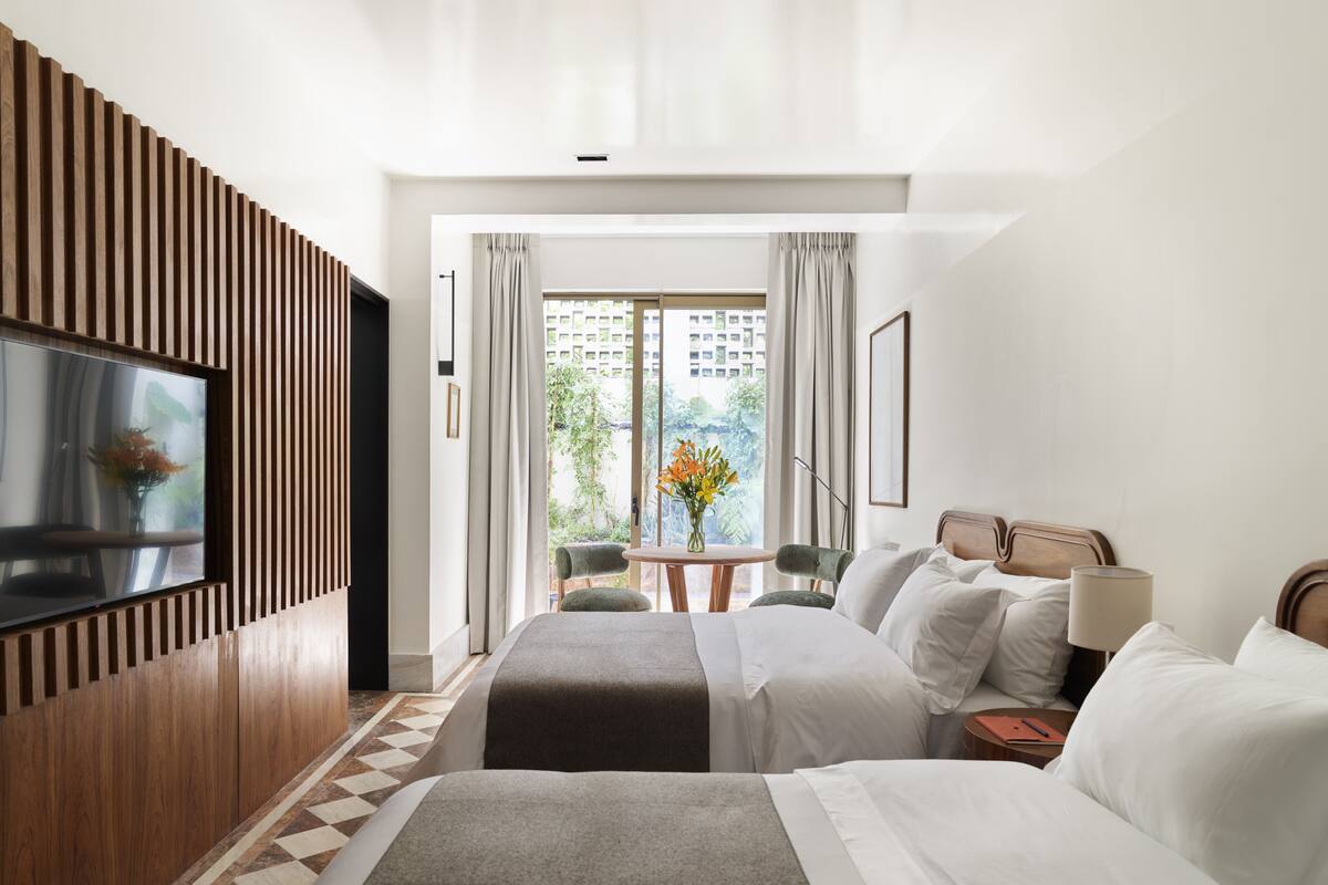 A hotel room with a large bed, a TV mounted on the wall, a small table with a vase of flowers, and a view of buildings through the window.