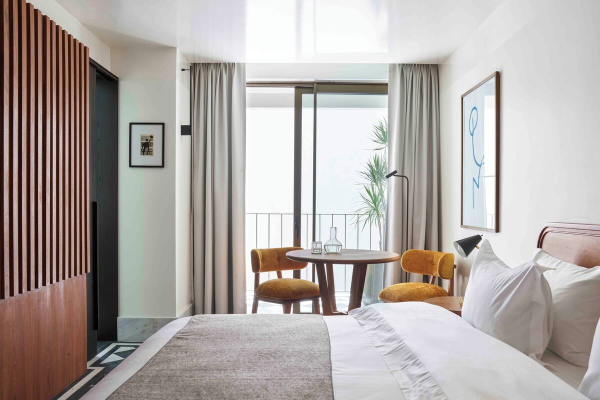 A hotel room with a large bed, a dining table with two chairs, a balcony with a view, and a framed artwork on the wall.
