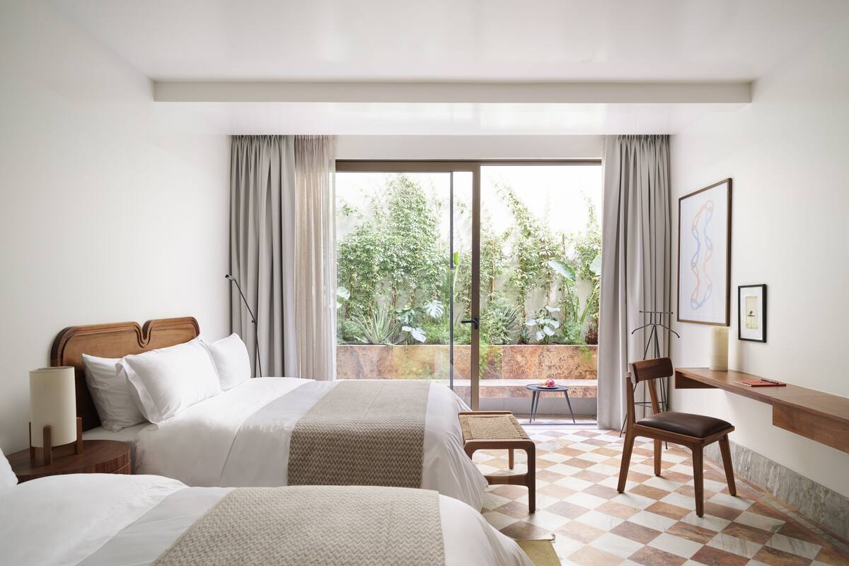 A hotel room with two beds, a desk, a chair, and a balcony with a view of greenery.