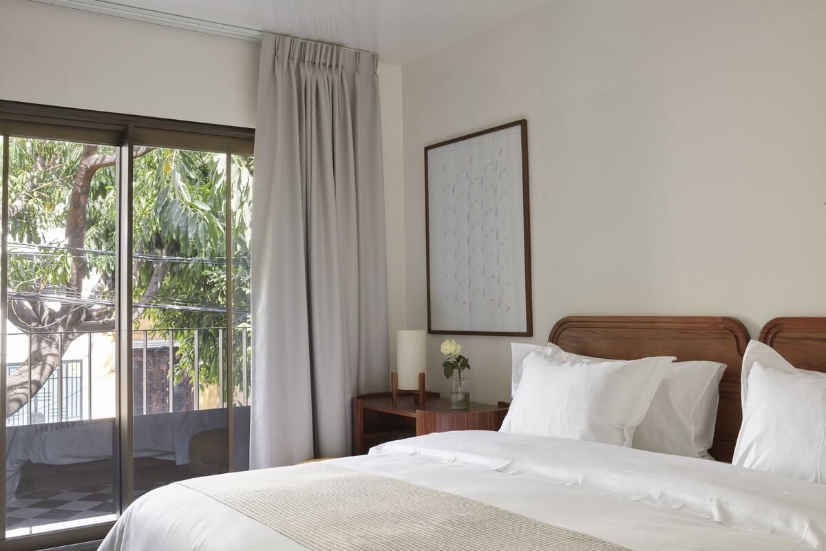 A hotel room with a large bed, a nightstand with a vase of flowers, a framed picture on the wall, and a view of greenery through the window.