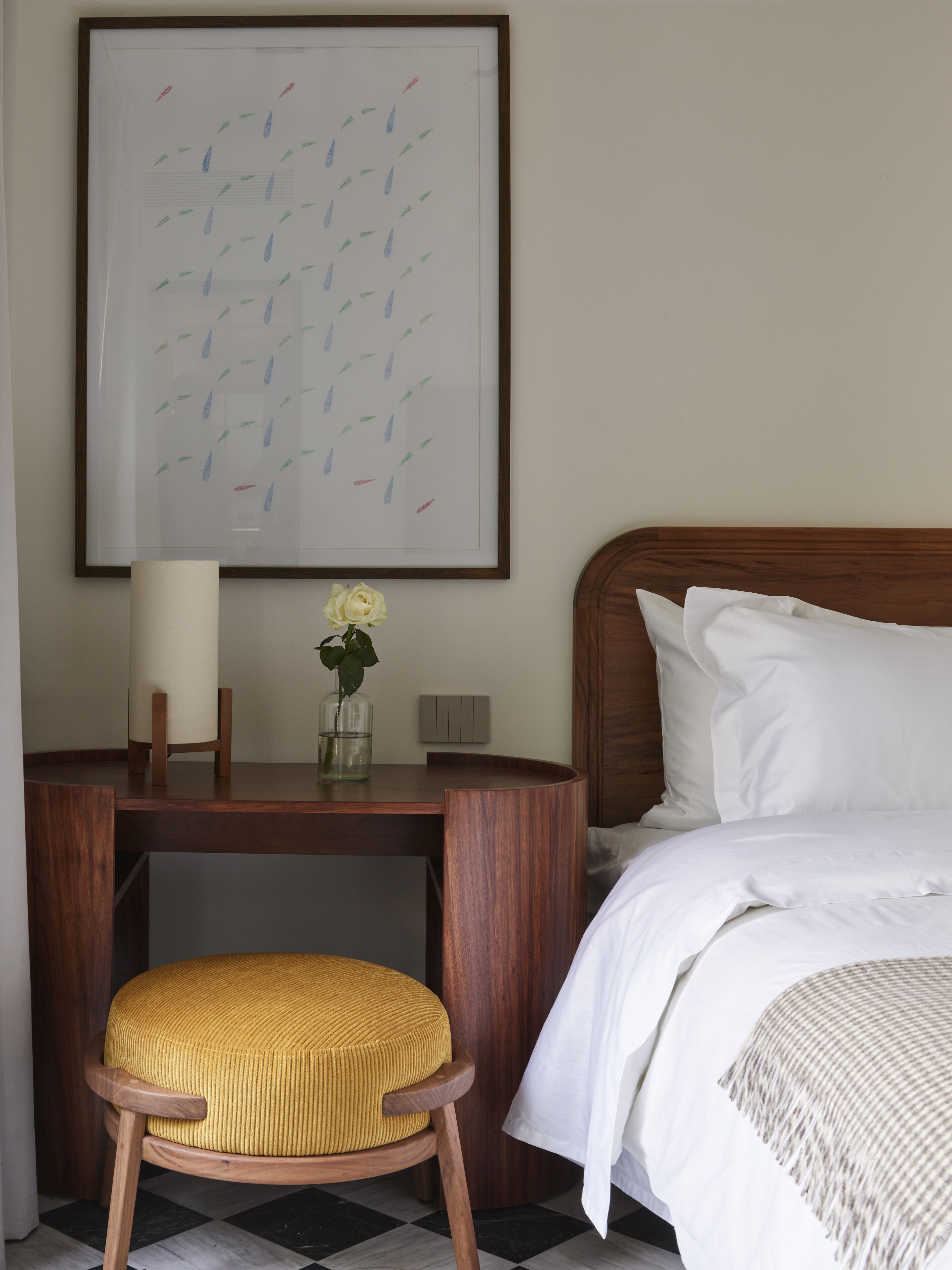 Een netjes ingerichte hotelkamer met een bed, een nachtkastje met een lamp en een bloemenvaas, een ingelijst kunstwerk aan de muur en een stoel met een kussen.