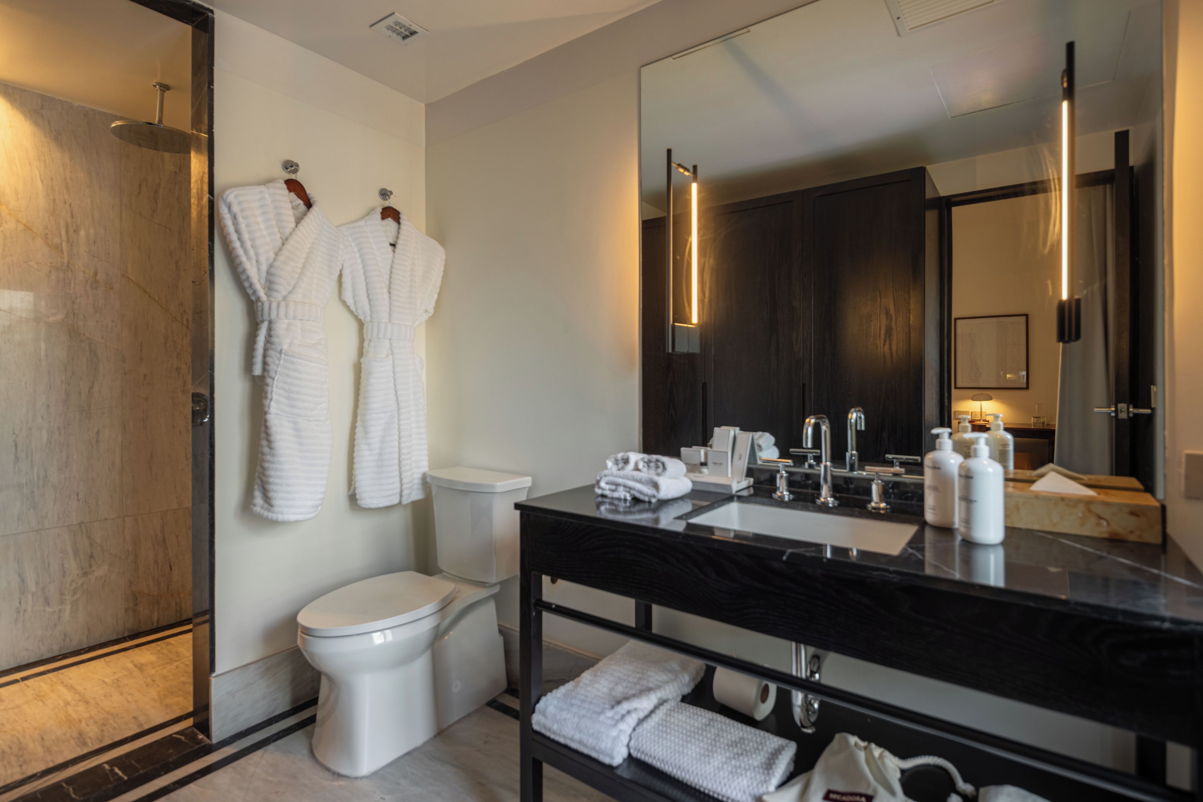 Un baño moderno con tocador de mármol negro, inodoro blanco y dos batas blancas colgadas en la pared.
