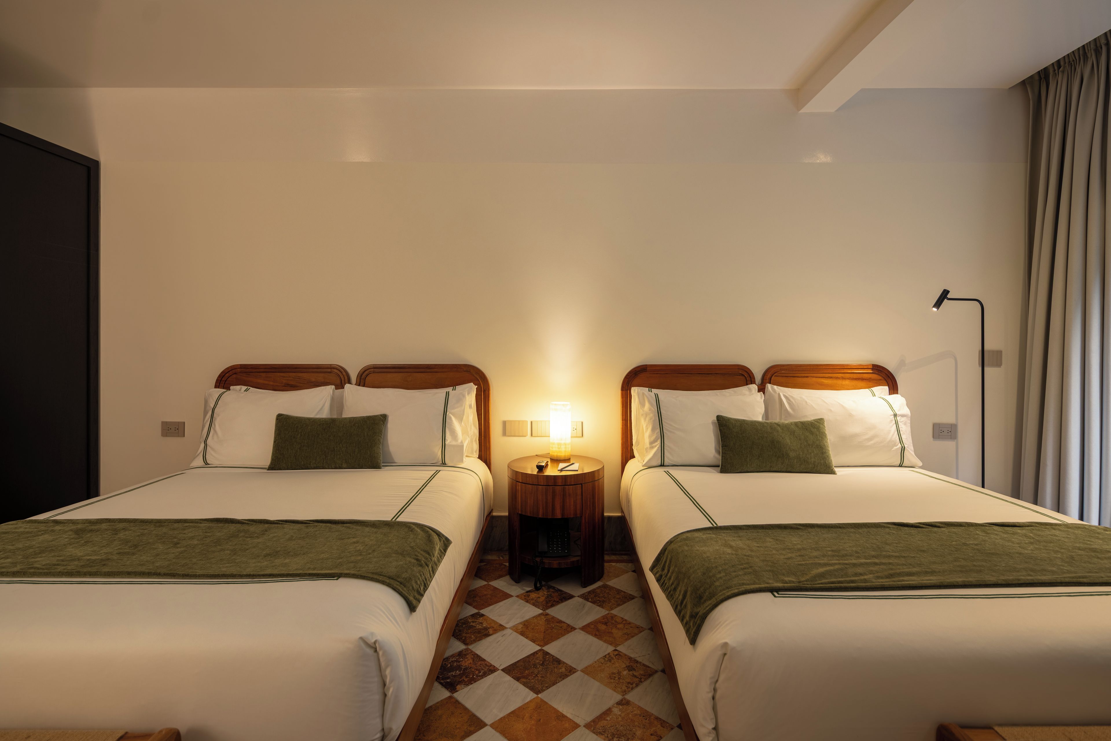 Een hotelkamer met twee bedden, een nachtkastje met lamp en een zwart-wit geruit vloerkleed.