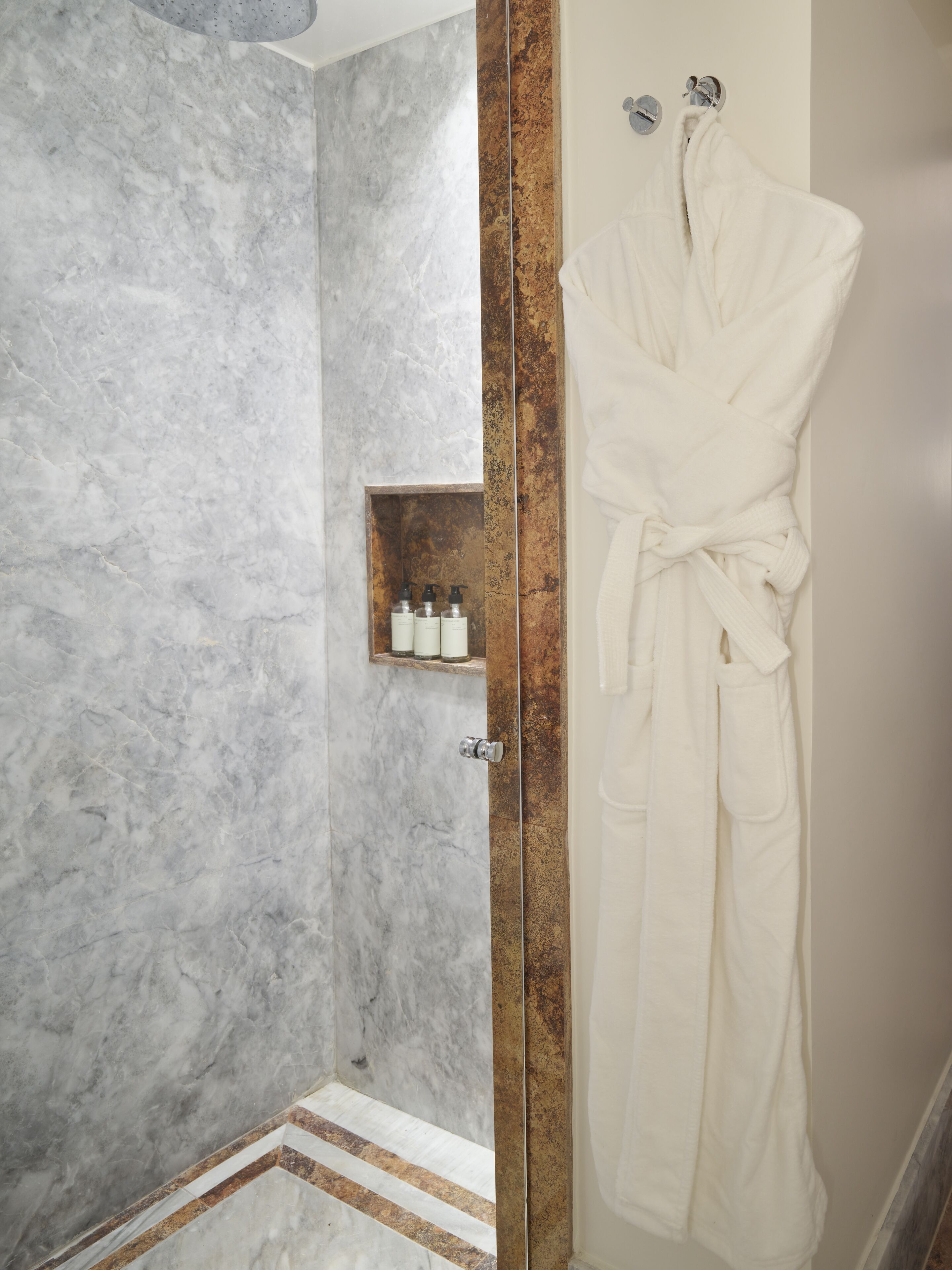 A bathroom with a marble wall, a glass shower enclosure, and a white robe hanging on a hook.