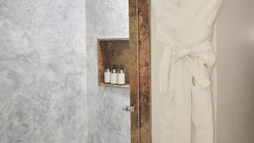 A bathroom with a marble wall, a glass shower enclosure, and a white robe hanging on a hook.