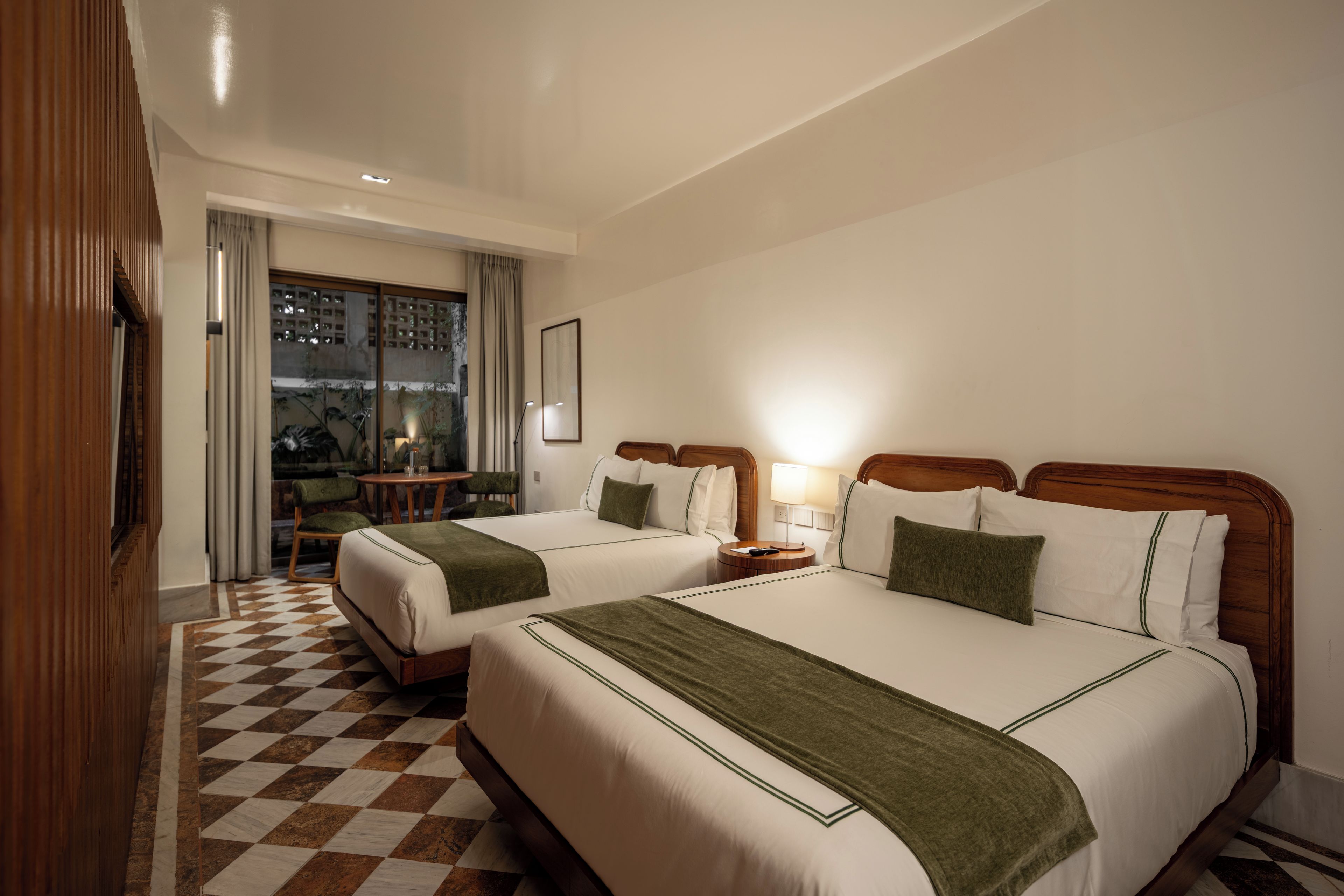 A hotel room with two beds, a checkered floor, a wooden headboard, a nightstand, a lamp, and a view of the outdoors.