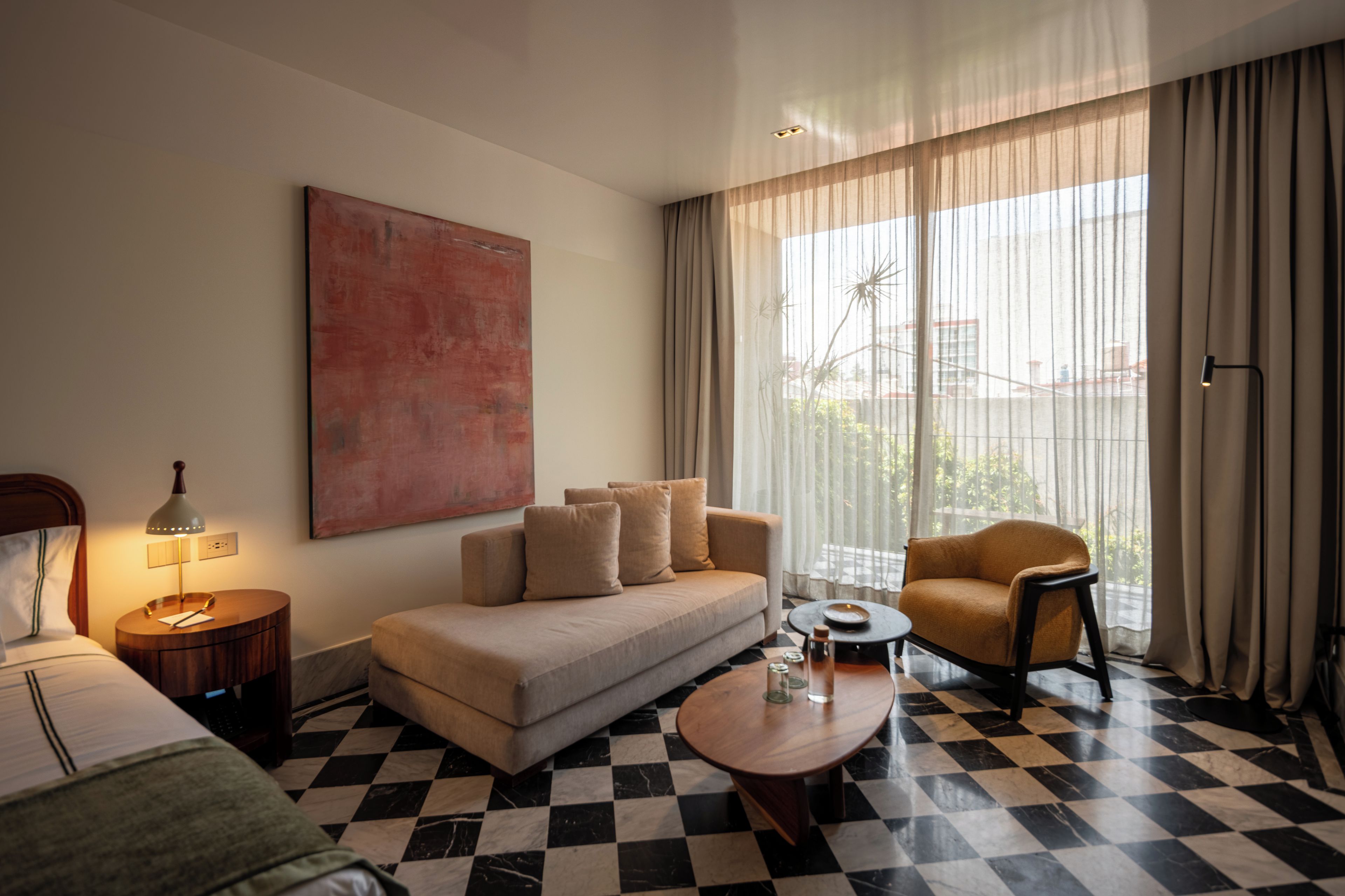 Habitación de hotel con piso a cuadros, un sofá, una silla, una mesa de centro, una lámpara, una cama y un ventanal con cortinas.