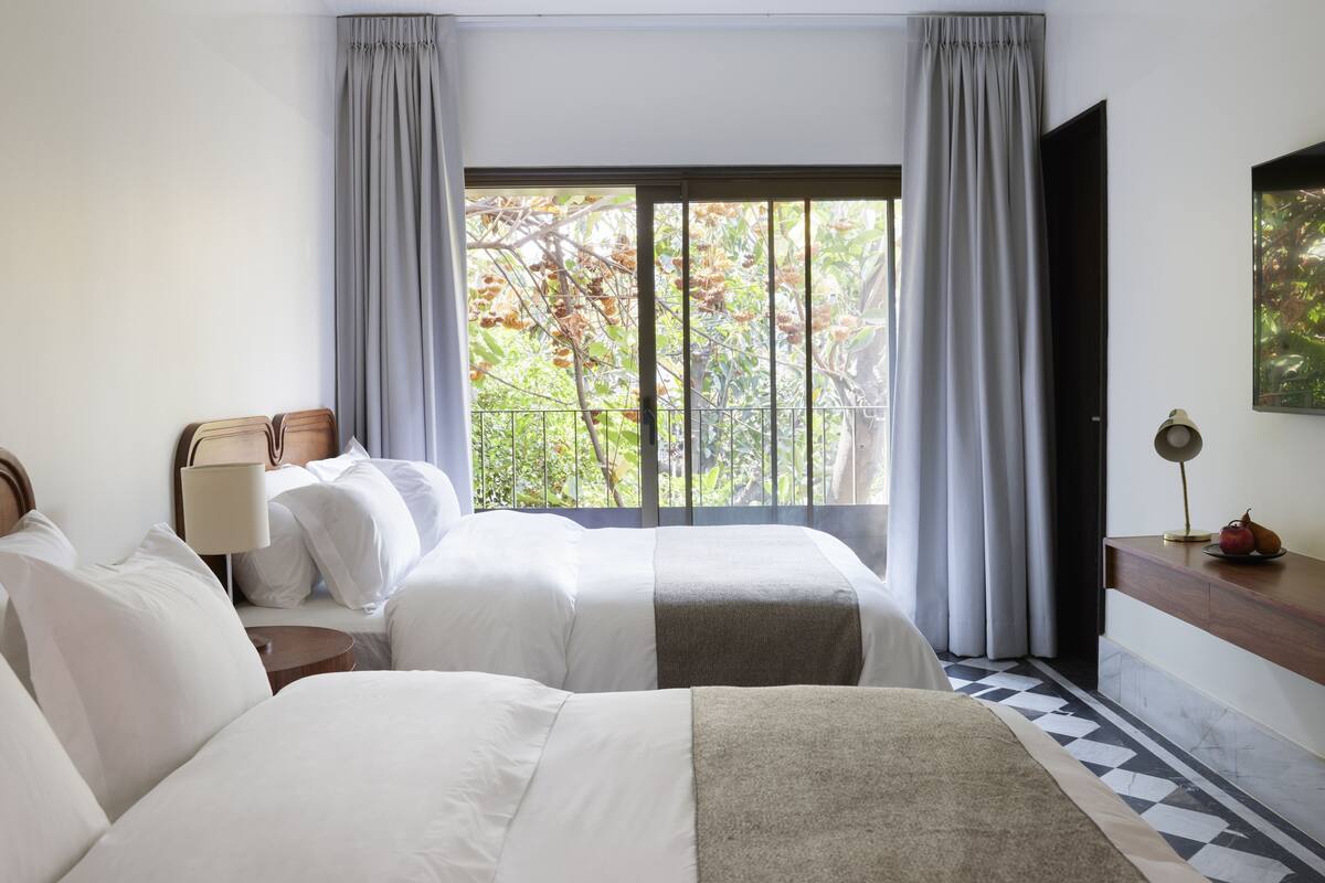 A hotel room with two beds, a desk, a lamp, and a window with a view of trees.