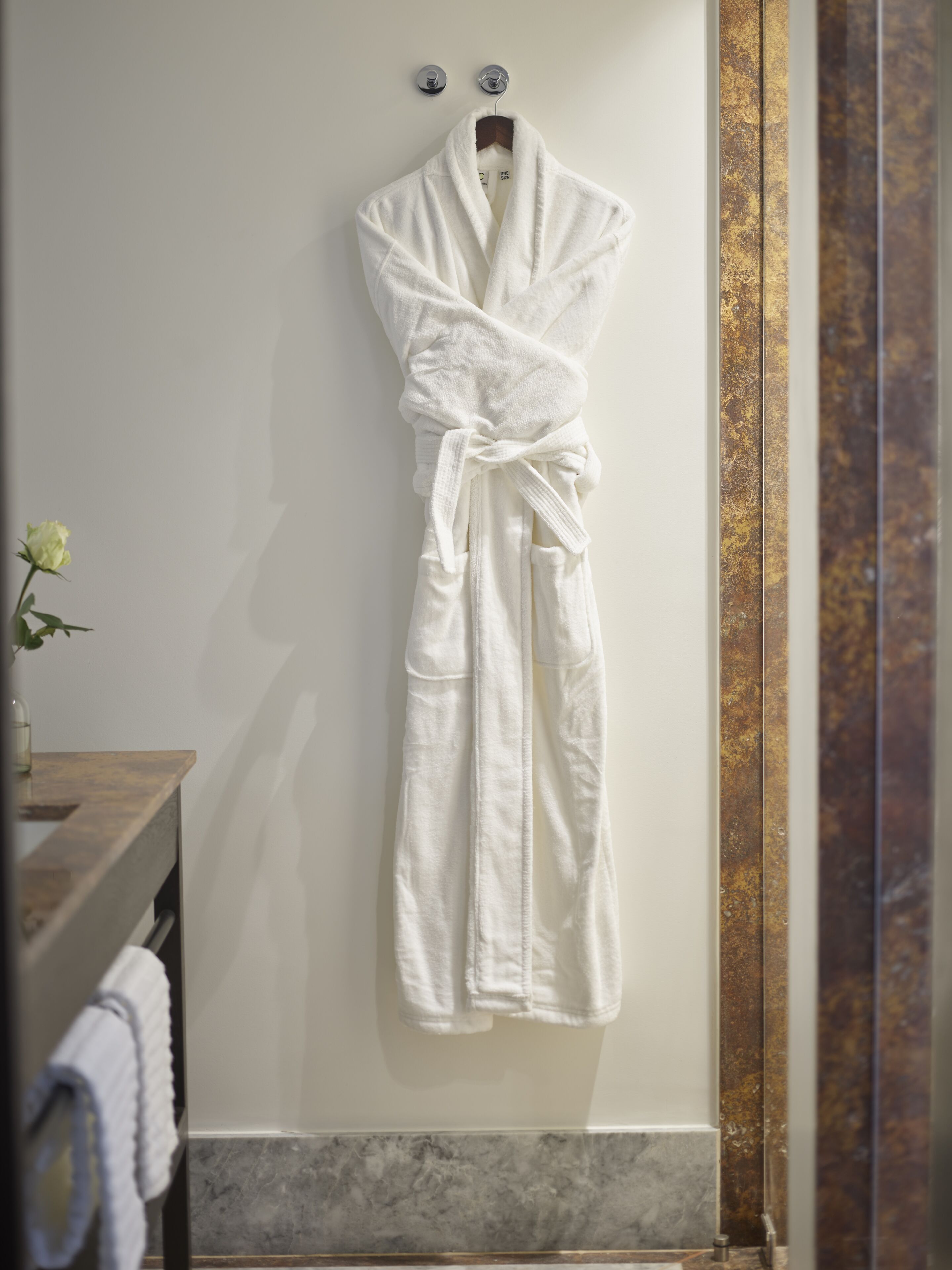 A white bathrobe hanging on a hook in a bathroom with a wooden vanity and marble flooring.