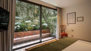 A room with a large sliding glass door leading to a garden, a small wooden table, a bench, and a framed artwork on the wall.