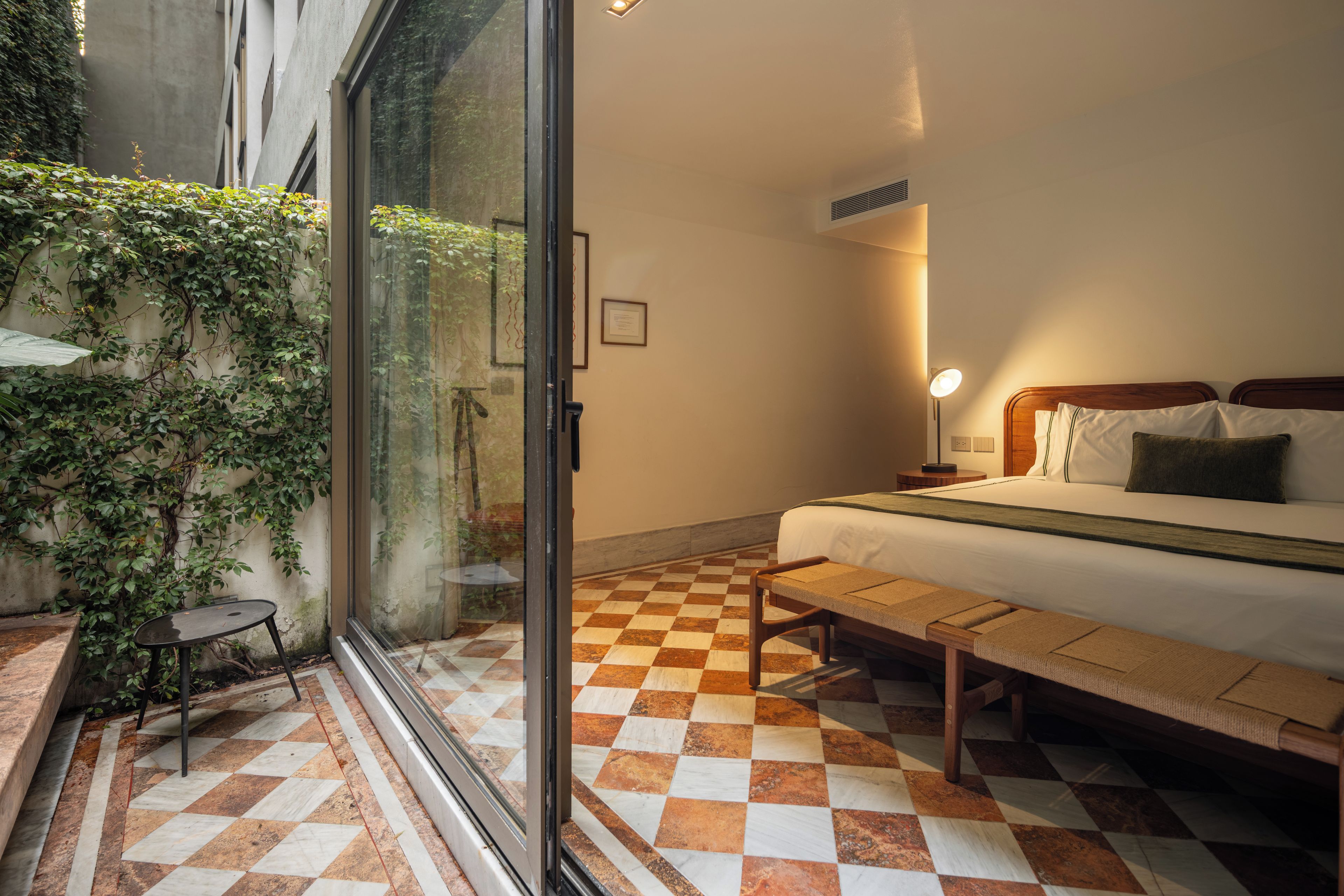 A hotel room with a checkered floor, a bed with a wooden headboard, a bedside table, a lamp, and a view of greenery through a glass door.