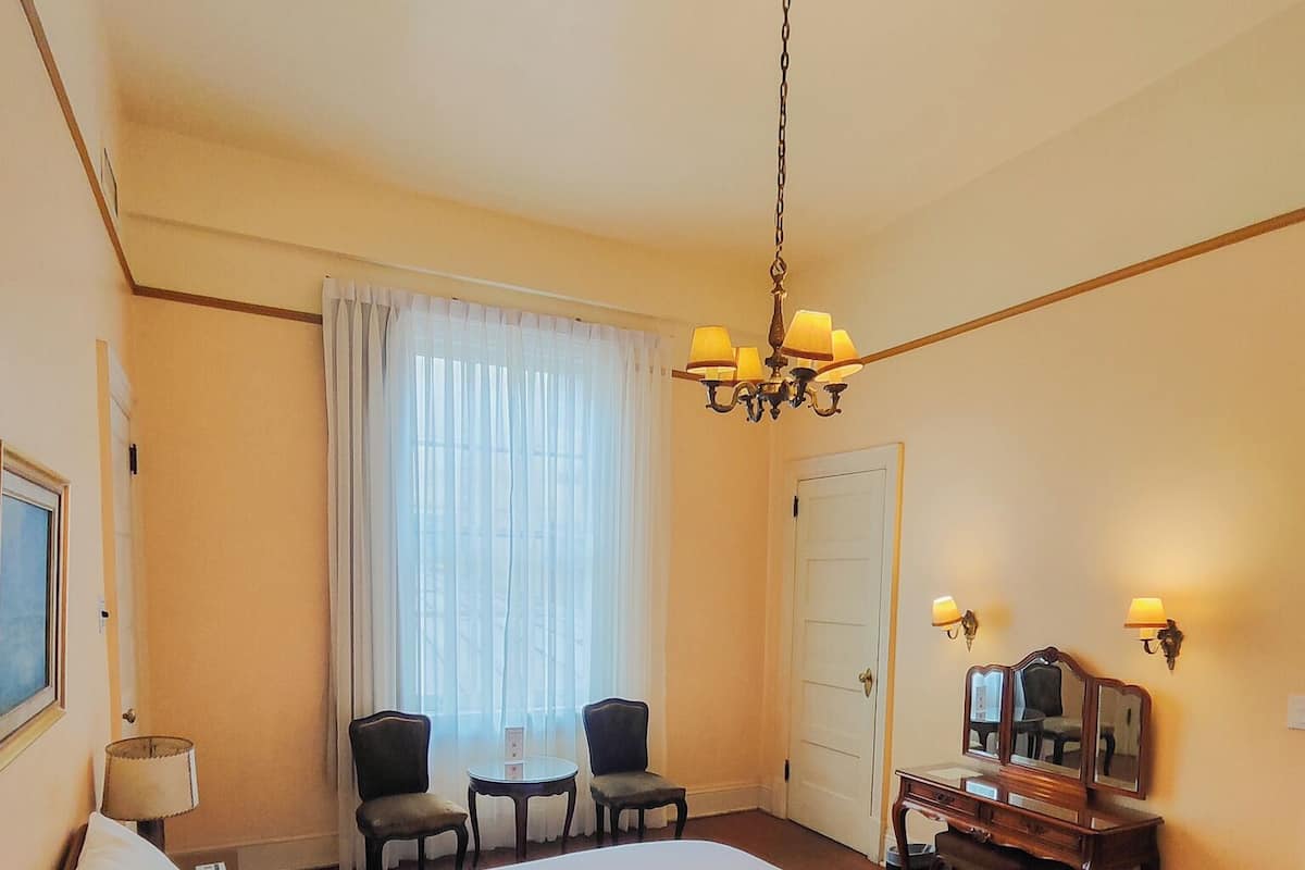 Una habitación de hotel con una cama grande, un escritorio con espejo, dos sillones, una araña y una ventana con cortinas.