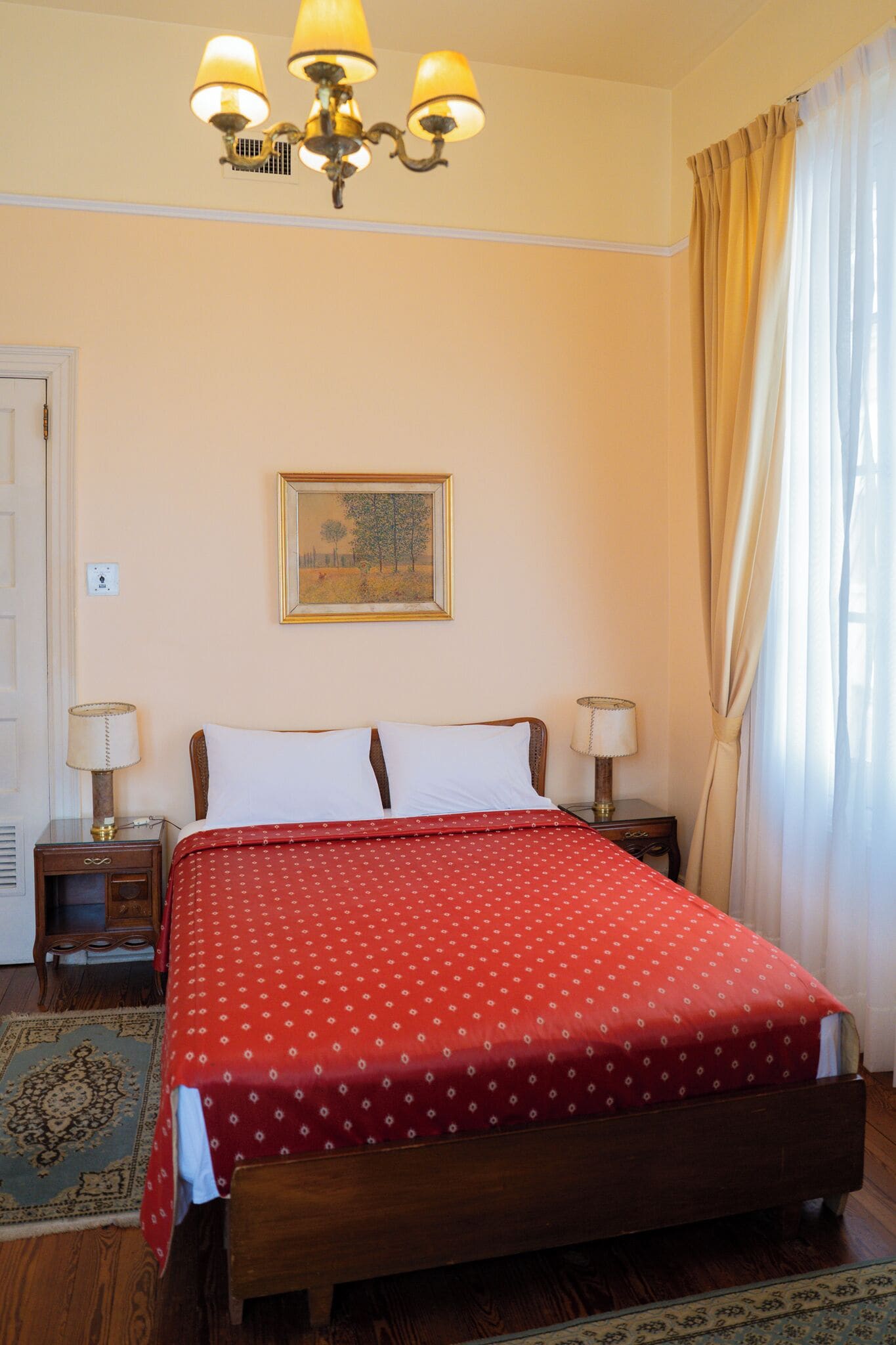 A bedroom with a wooden bed, two bedside tables with lamps, a chandelier, a framed picture on the wall, and a window with curtains.