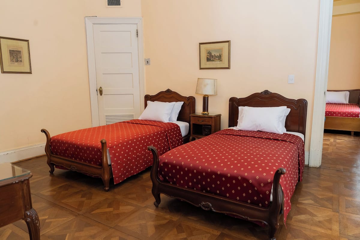 Una habitación con dos camas de madera, una mesita de noche con lámpara, una puerta y un cuadro enmarcado en la pared.