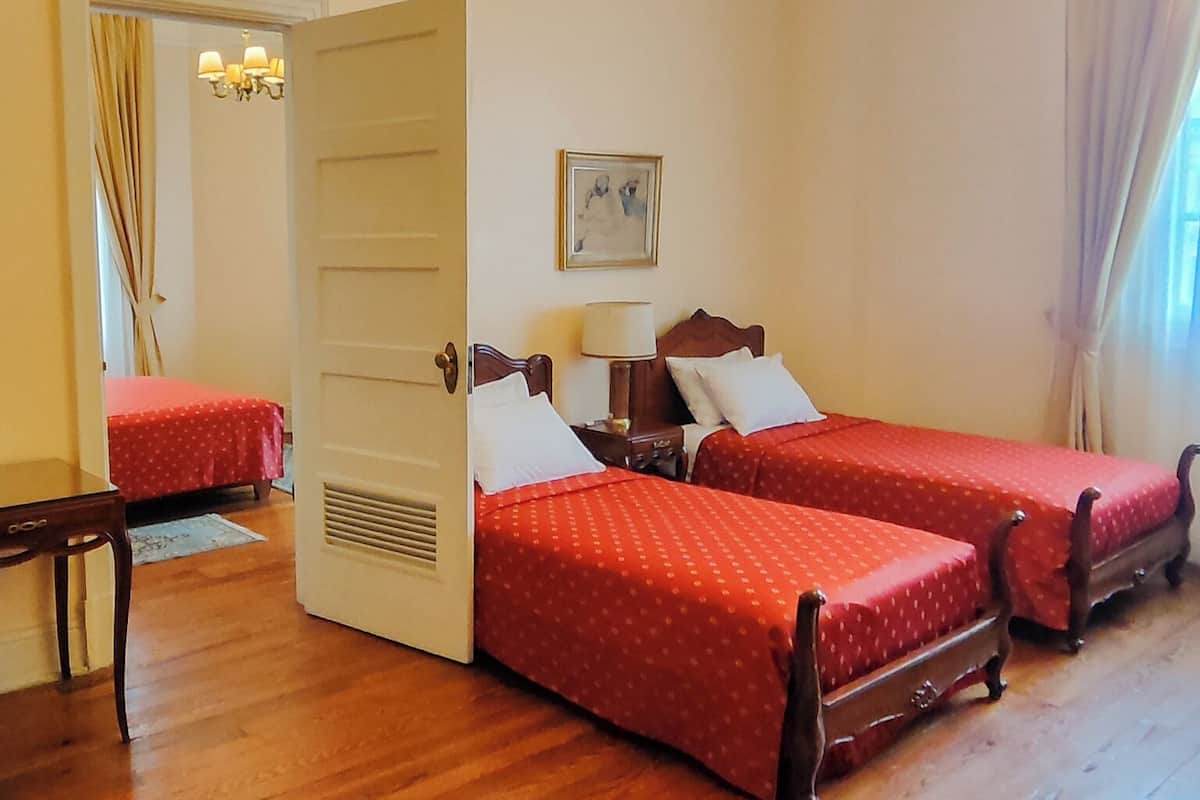 Habitación de hotel con dos camas, piso de madera, candelabro, puerta y un cuadro en la pared.
