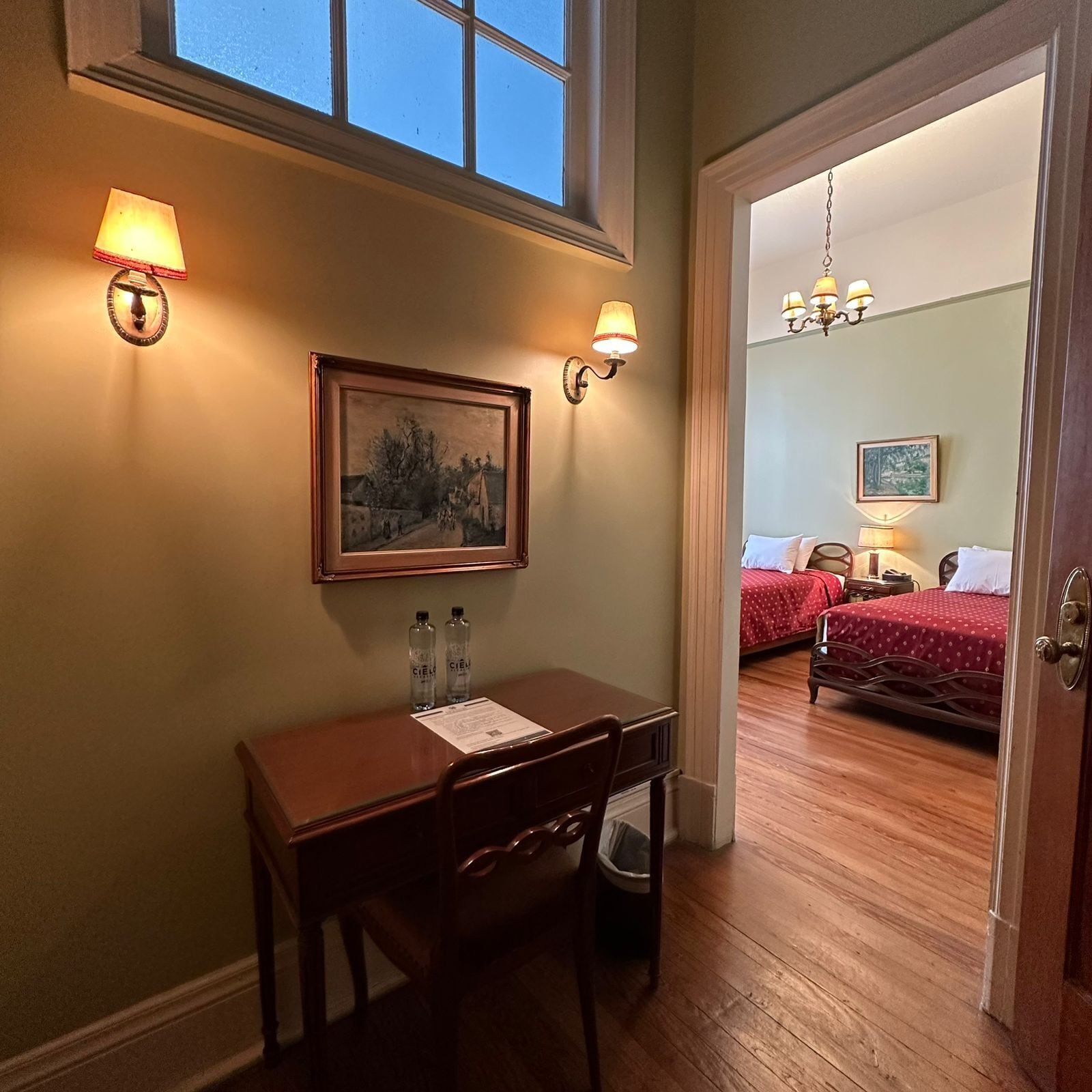Een hotelkamer met een bureau, stoel en een schilderij aan de muur. Er is een bed met een rode sprei en een kroonluchter in de hoek.