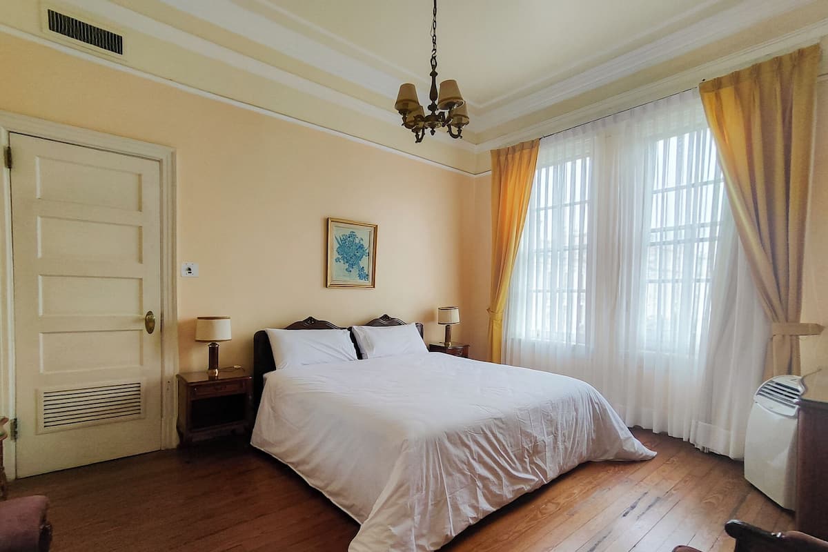 Una habitación de hotel con una cama grande, dos mesitas de noche, un ventilador de techo, una ventana con cortinas y una puerta.