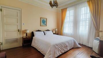 Una habitación de hotel con una cama grande, dos mesitas de noche, un ventilador de techo, una ventana con cortinas y una puerta.