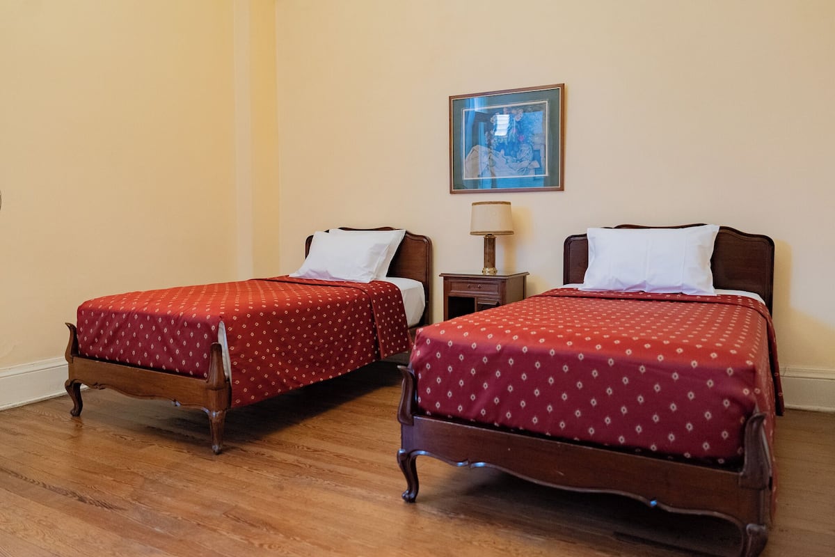 Dos camas de madera con ropa de cama estampada roja, una mesita de noche con una lámpara y un cuadro en la pared.