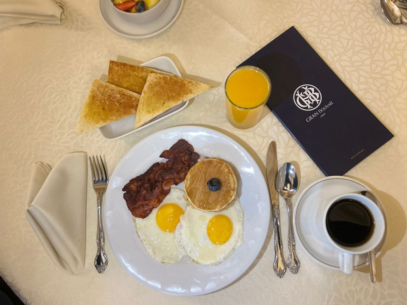 A breakfast plate with eggs, bacon, and a blueberry, toast, a glass of orange juice, and a menu.