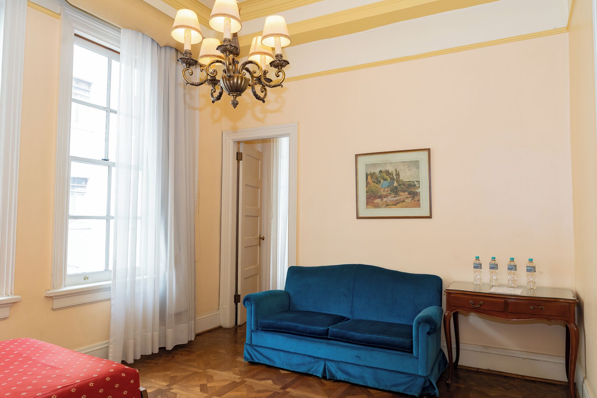 A room with a blue sofa, a wooden side table with water bottles, a framed picture on the wall, and a window with white curtains.