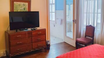 Ein Zimmer mit einem hölzernen Schminktisch, einem Fernseher, einem Bett mit roter Decke, einem Stuhl und einer Tür zu einem Außenbereich.