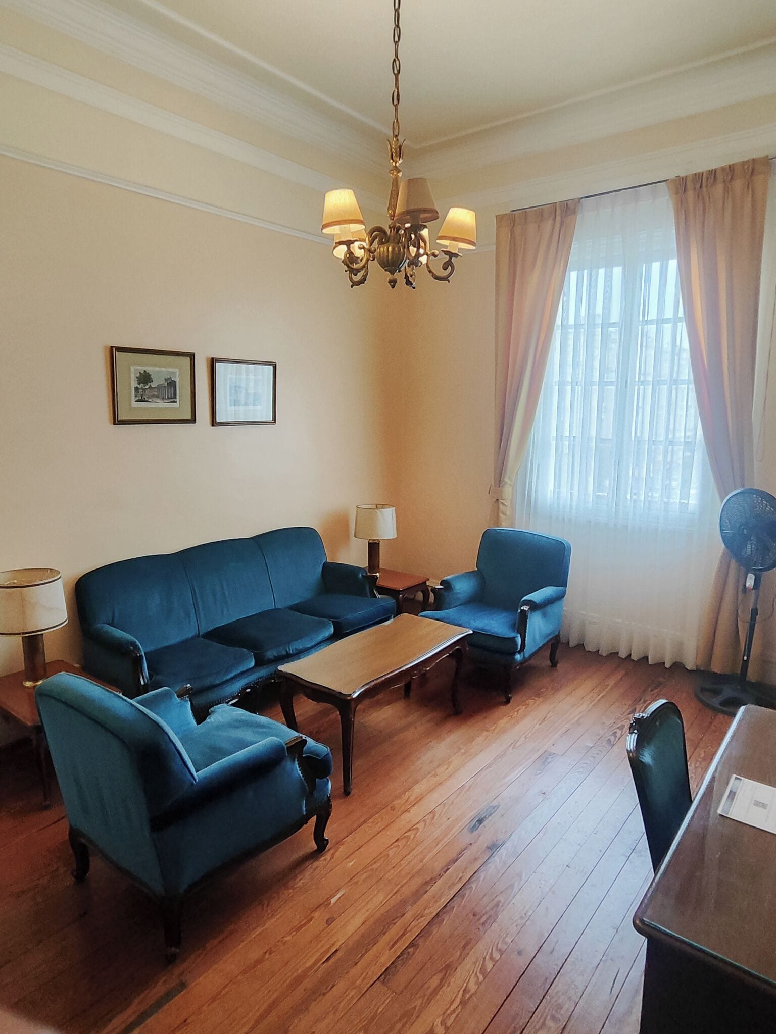 Ein Wohnzimmer mit einer Couch, zwei Sesseln, einem Couchtisch, einer Pendelleuchte, einem Ventilator und zwei gerahmten Bildern an der Wand.