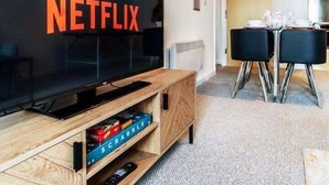 A living room with a large flat-screen TV displaying "NETFLIX" on the screen, a wooden entertainment unit with books and a remote control, a dining area with a table and chairs, and a carpeted floor. - Aspire City Centre Suite - PingPong & Netflix (West Midlands)