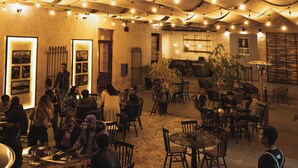 Un área de comedor al aire libre con mesas y sillas bajo toldos blancos, luces colgantes y piso de piedra.