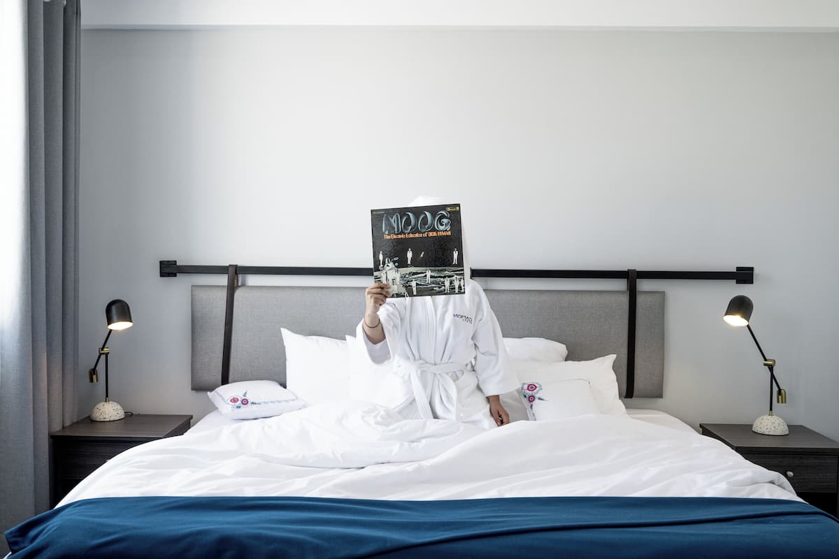 Una habitació d'hotel amb un llit, dues làmpades de nit, una taula de nit, una bata blanca i un llibre anomenat "MOOG" sobre el llit.