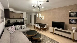 Una sala de estar moderna con un sofá gris, una mesa de comedor con sillas, un televisor sobre un mueble bajo y una cocina al fondo.
