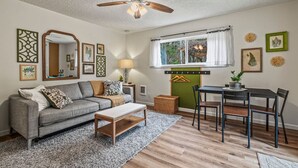 A living room with a sofa, coffee table, and dining table with chairs. There is a ceiling fan, a window with curtains, and wall decorations. - Waterfront Getaway w/ Sauna and BBQ (Longview)