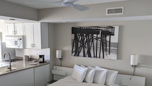 A modern hotel room with a large bed, a kitchenette, a ceiling fan, and a painting of a pier. - Harbor Inn (Wrightsville Beach)