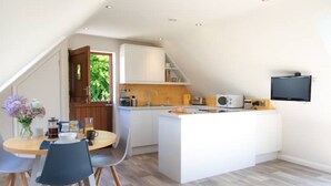 A modern kitchen with a central island, a dining area with chairs, and a TV mounted on the wall. - The Roost in the Blackdowns (Hemyock)