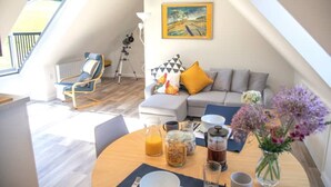 A cozy living area with a sofa, a dining table with breakfast items, a chair, and a painting on the wall. - The Roost in the Blackdowns (Hemyock)