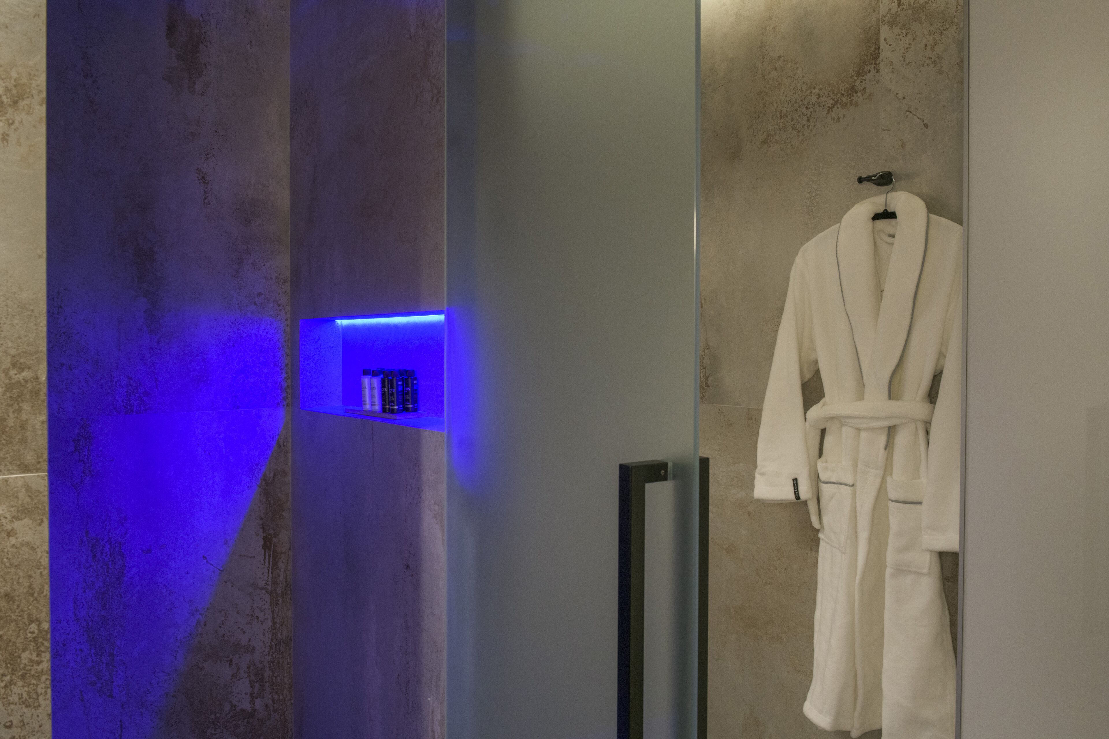 Un bagno con un accappatoio bianco appeso a un gancio, un pannello illuminato di blu e un portaservizi.