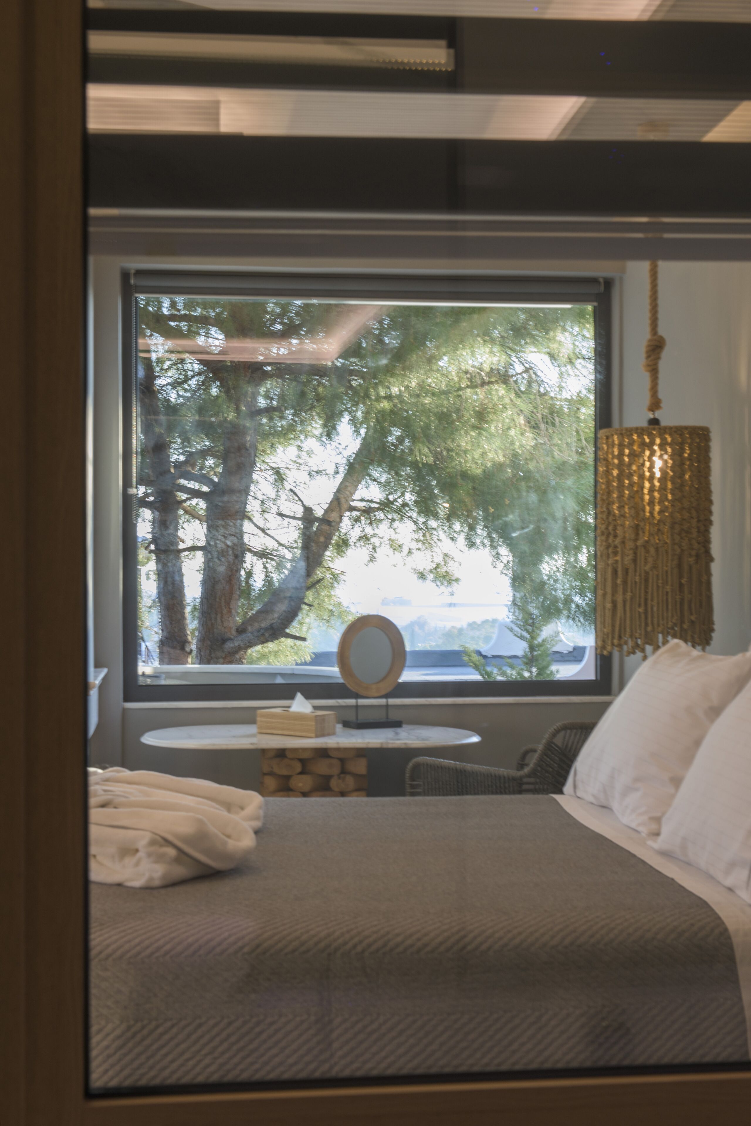 A bedroom with a large window, a bed, a bedside table, a hanging lamp, and a view of trees outside.