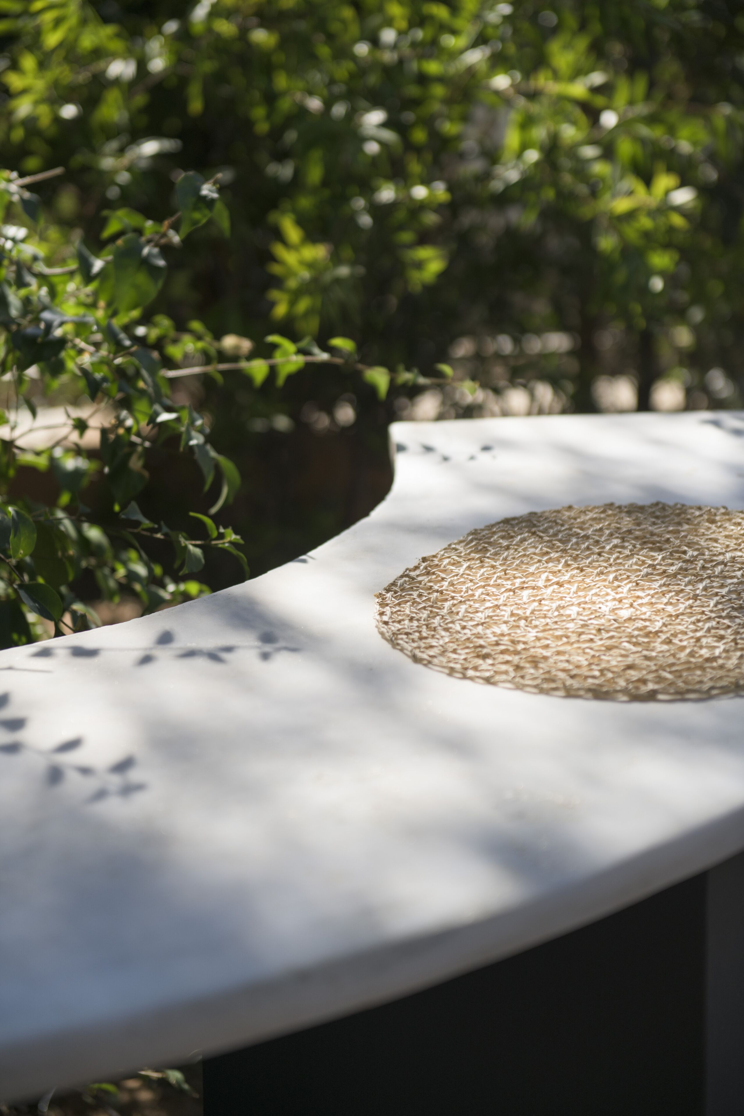 A white outdoor table with a woven mat on top, surrounded by greenery.