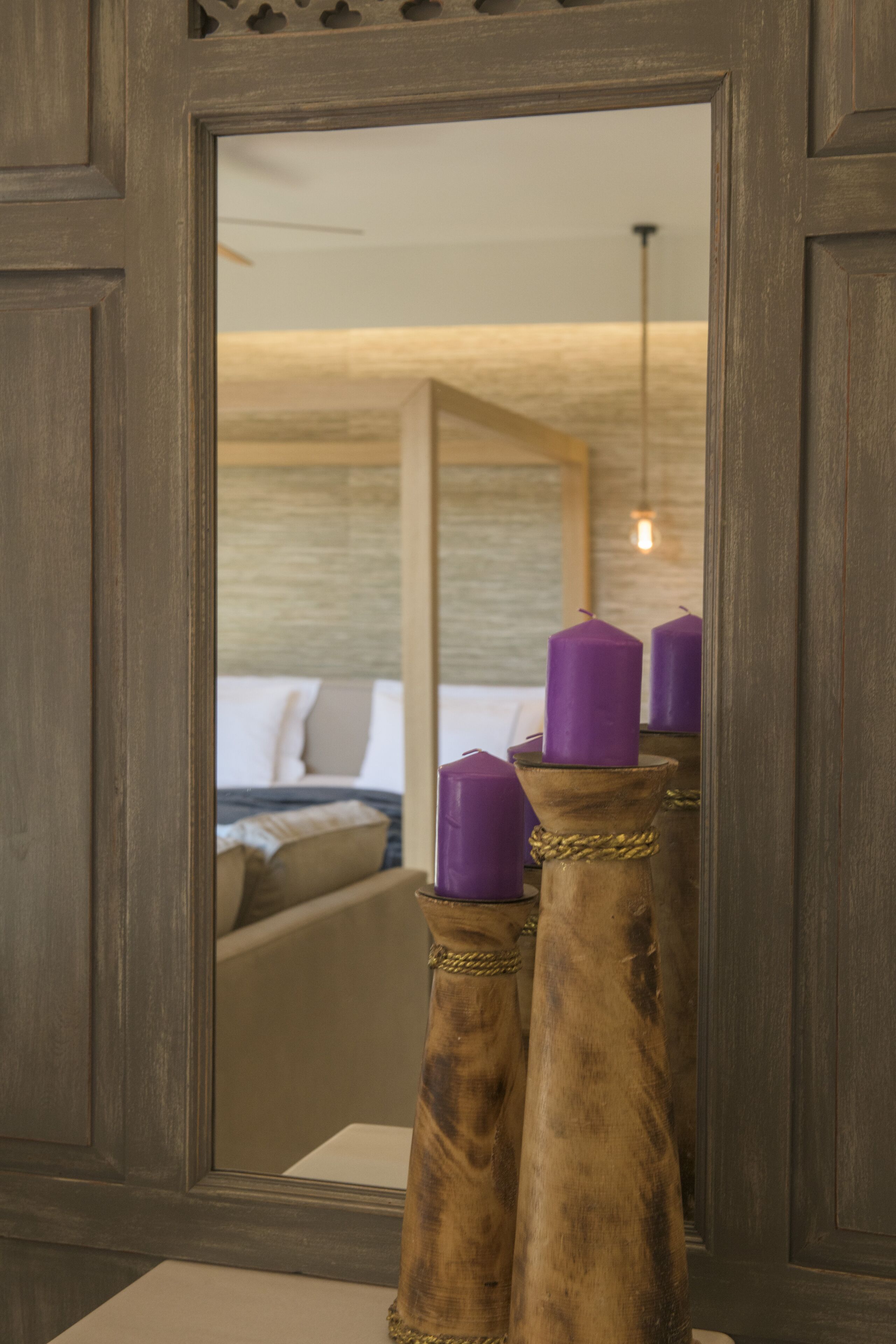 A mirror reflecting a room with a bed, a hanging light fixture, and a wooden object with purple candles.