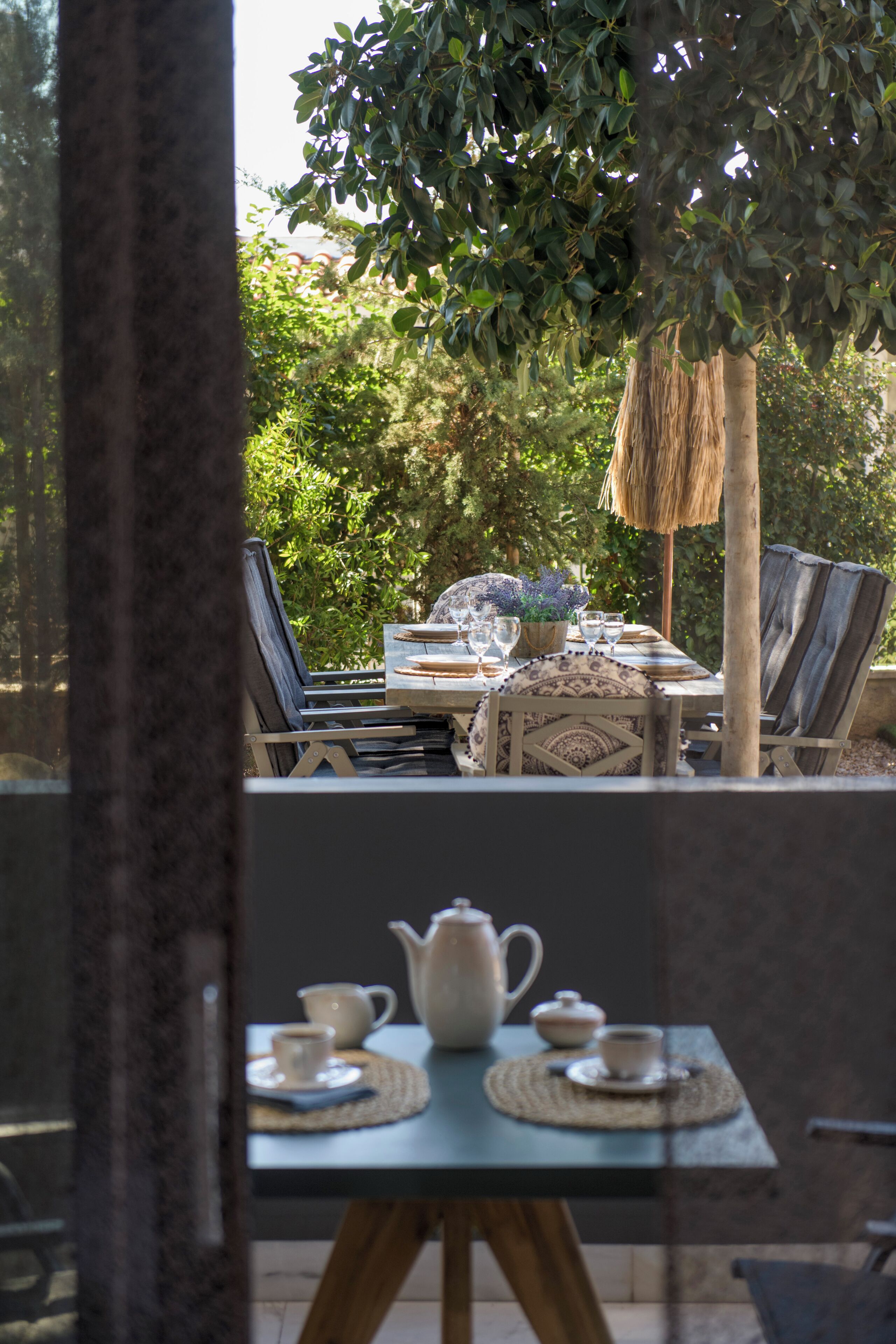 An outdoor dining area with a table set for two, surrounded by chairs, a thatched umbrella, and lush greenery in the background.