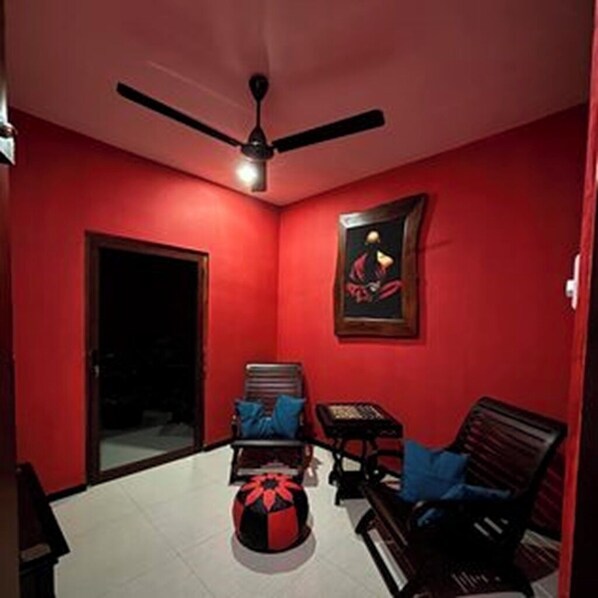 Une pièce aux murs rouges, avec un ventilateur au plafond, un tableau encadré, une table entourée de chaises et un petit objet décoratif posé sur le sol.