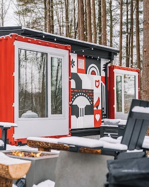 Une cabane dotée de grandes fenêtres, d’un foyer extérieur et d’une terrasse.