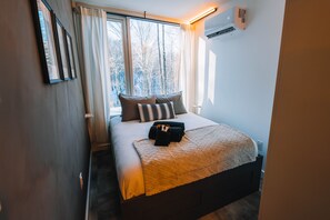 A neatly made bed with a quilted blanket, two pillows, and a set of towels. There is a window with curtains, an air conditioning unit, and a framed picture on the wall.